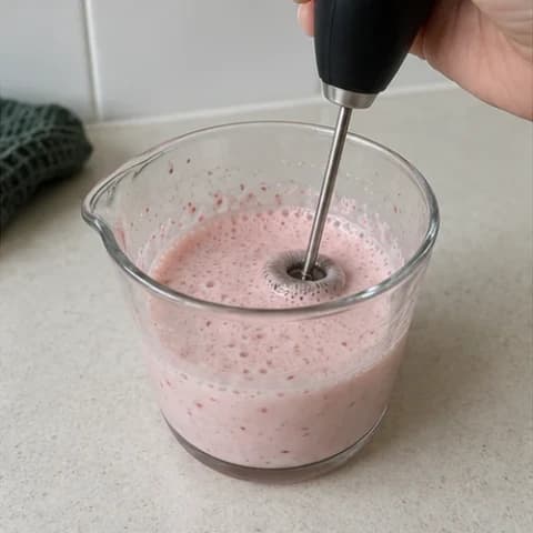 A handheld frother mixing the pink strawberry cream in a glass cup