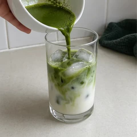 Whisked matcha being poured into a clear glass of milk and ice