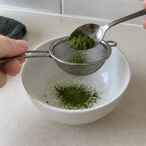 Matcha powder being sifted into a white bowl through a fine metal sieve