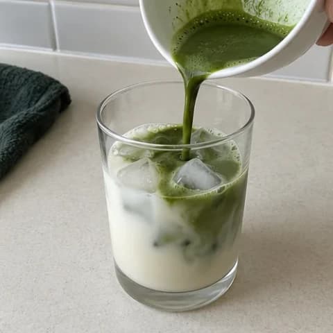 Green matcha being poured into a clear glass filled with milk and ice