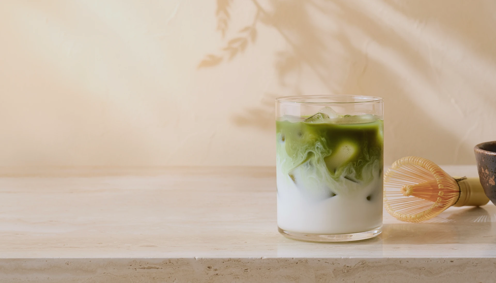 Iced matcha and milk in a clear glass with a bamboo whisk on pale stone